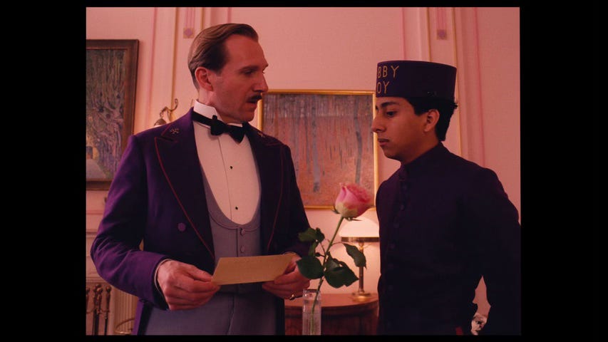 Still from The Grand Budapest Hotel. M. Gustave explains the content of a telegram to Zero in a pink hotel room with art on the walls. A single rose in a vase sits on a table between them.