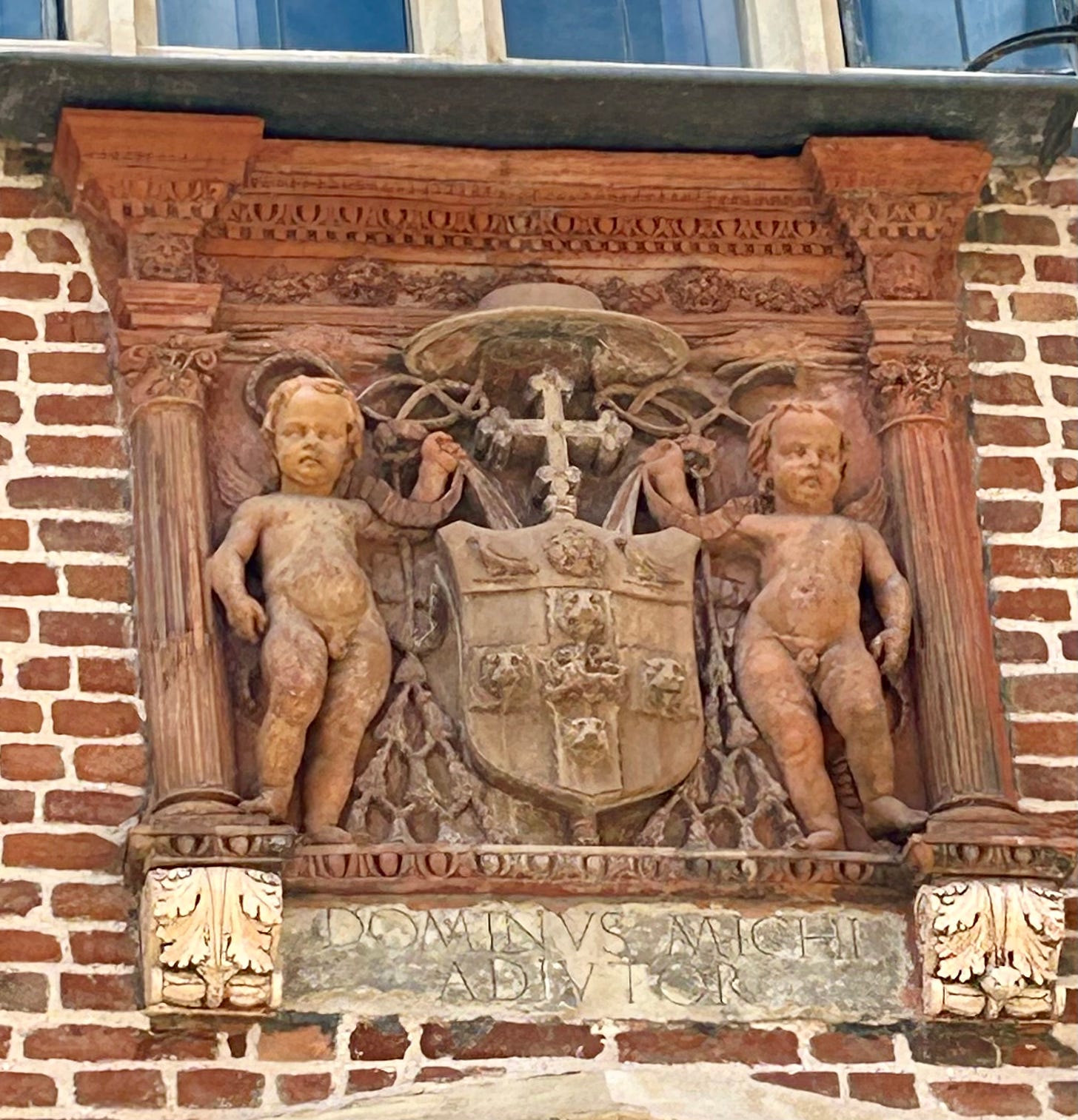 Brickwork including a coat of arms featuring a cardinal’s hat and two birds, Tudor rose and lions’ heads. Two putti are on either side of the arms. 