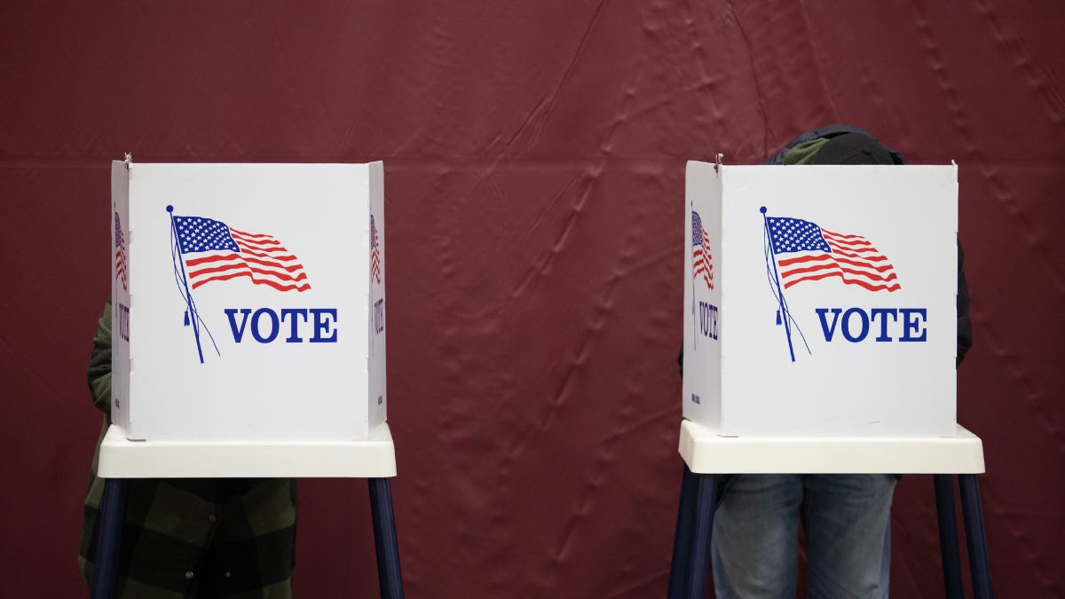 Two white voting booths with bold "VOTE" text and an illustration of the American flag on them. Individuals are seen from behind, partially obscured as they cast their votes, against a maroon background. Two white voting booths with bold "VOTE" text and an illustration of the American flag on them. Individuals are seen from behind, partially obscured as they cast their votes, against a maroon background.