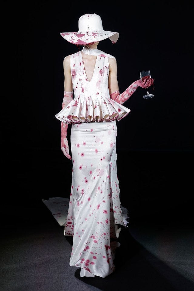 a high fashion model dressed in Robert Wun's debut couture collection; she wears a hat, gown, and gloves that appear to be wine-stained while holding a wine glass full of beads.