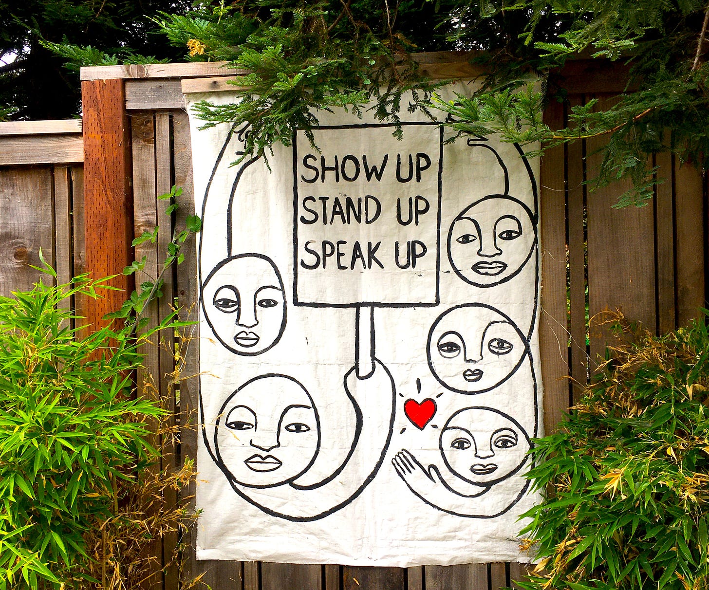 a hand painted sign surrounded by trees and plants. It shows five line-drawing faces and the words "Show Up. Stand Up. Speak Up." plus a red heart