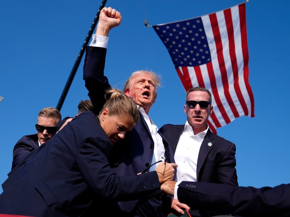 A man raises his fist with blood smeared across his face after being hit with a bullet on his ear with an American flag in the background.