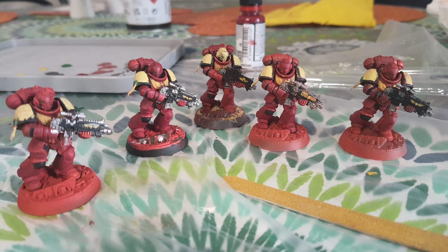 The picture shows a set of Space Marine miniatures from Warhammer 40k, painted in the red colour pattern of the Blood Ravens chapter (that is, space soldiers with power armuor and weapons). In the background, various paints and painting implements.