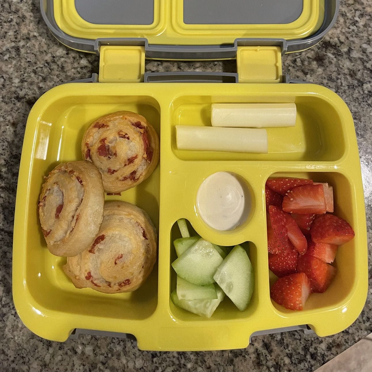 An open lunchbox with pizza pinwheeels, string cheese, strawberries, cucumbers and ranch. An open lunchbox with pizza pinwheeels, string cheese, strawberries, cucumbers and ranch.