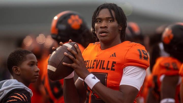 A young football player in an orange jersey holds a football, looking focused. A younger boy stands nearby, watching.