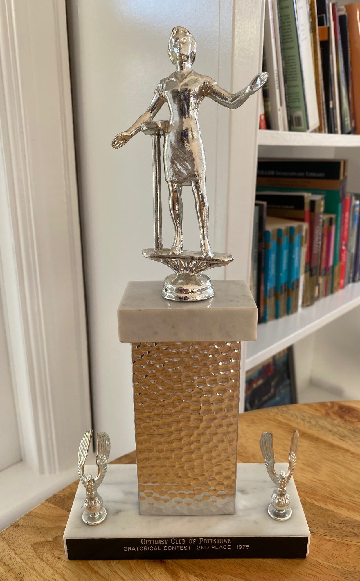 Photo of a trophy on a table with bookshelves in the background. A female figure with an outstretched left arm is atop the trophy and the inscription below reads: "Optimist Club of Pottstown/Oratorical contest 2nd place 1975"