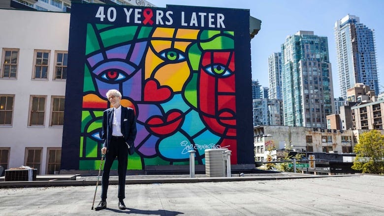 A man stands in front of a large colourful mural with skyscrapers in the background. 