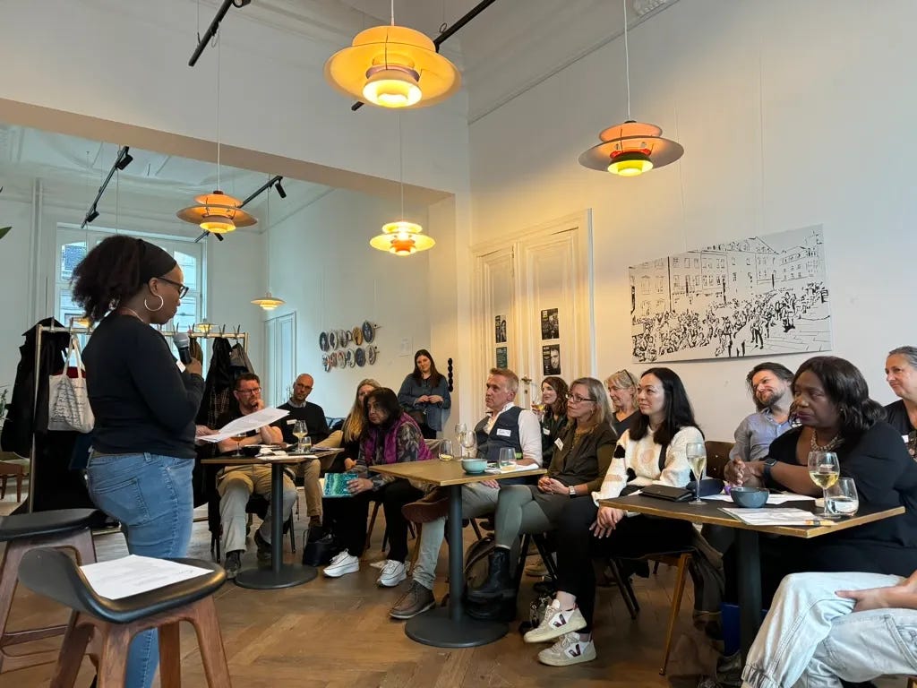 A woman reads her work to a room of people sitting at small tables. The room is brigh and has midcentury chandeliers