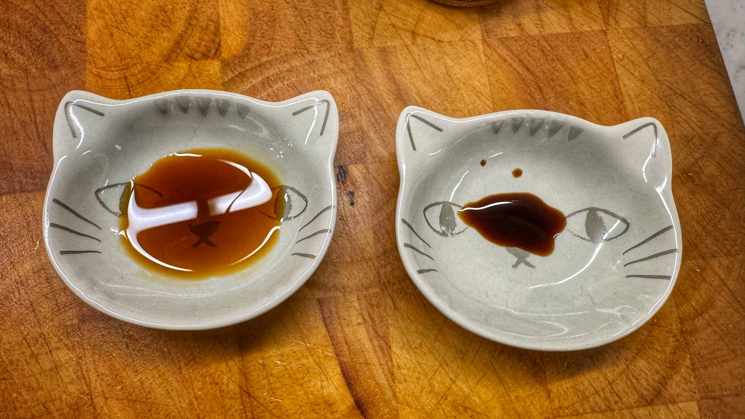 Two shallow dishes shaped like cats faces are on a cutting board. Both contain small amounts of soy sauce. The sauce on the left is medium brown and the bottom of the bowl is visible. The sauce on the right is very dark brown can cannot be seen through.