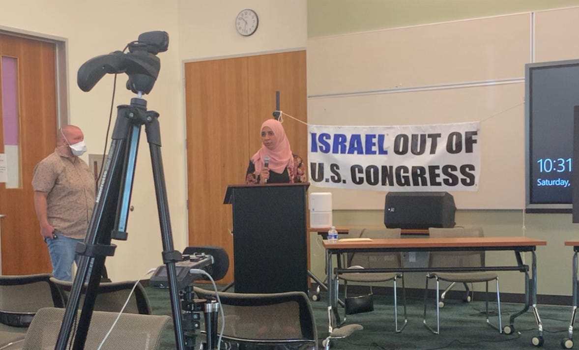 A woman wearing a pink hijab speaks into a microphone in front of
a podium, a banner reading Israel Out of Congress behind her.
