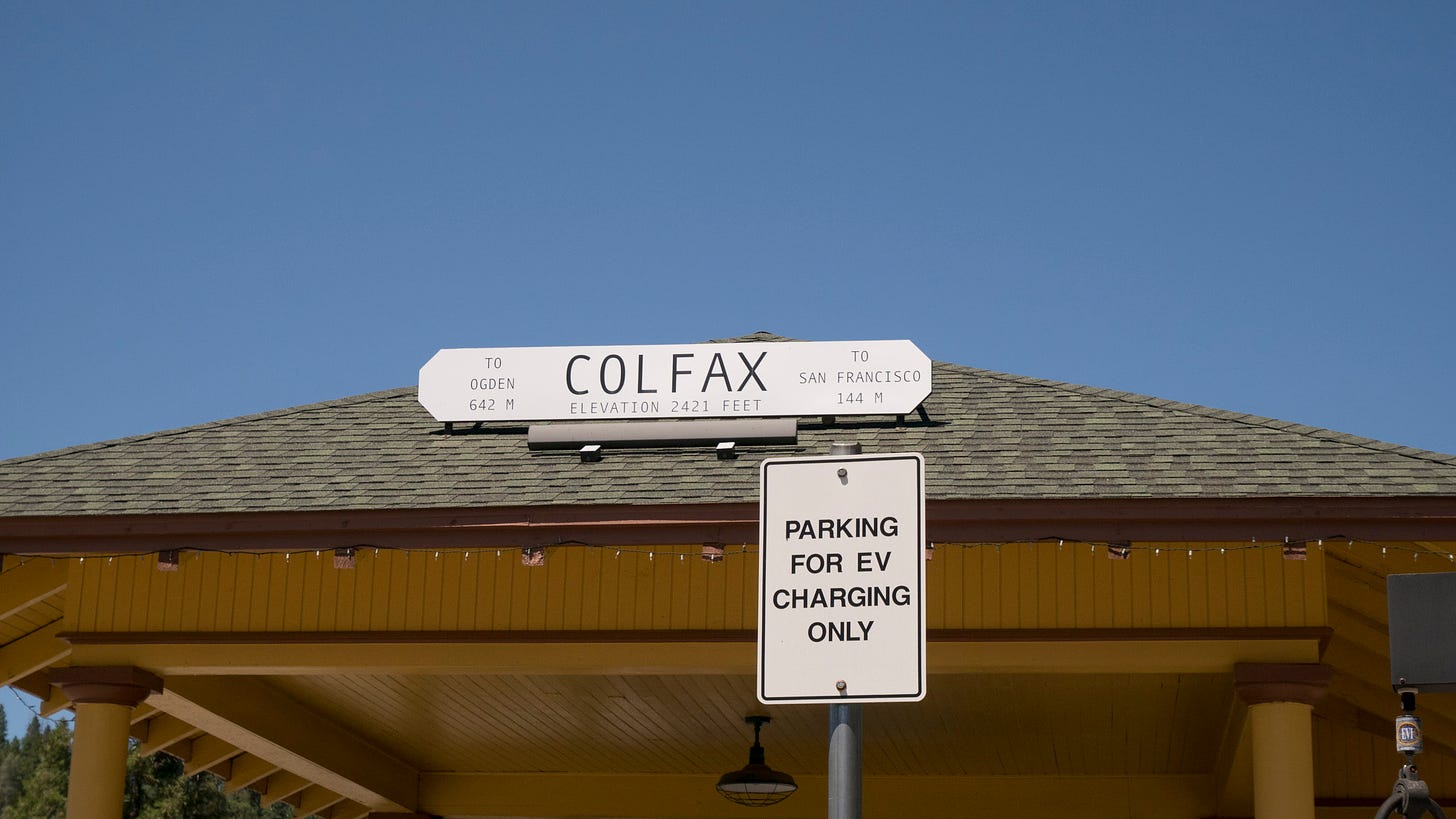 The peaked roof of an old railroad station building. A simple wooden sign on the roof says, "COLFAX ELEVATION 2421 FEET. TO OGDEN 642 M. TO SAN FRANCISCO 144 M". In front, another sign on a post says, "PARKING FOR EV CHARGING ONLY"