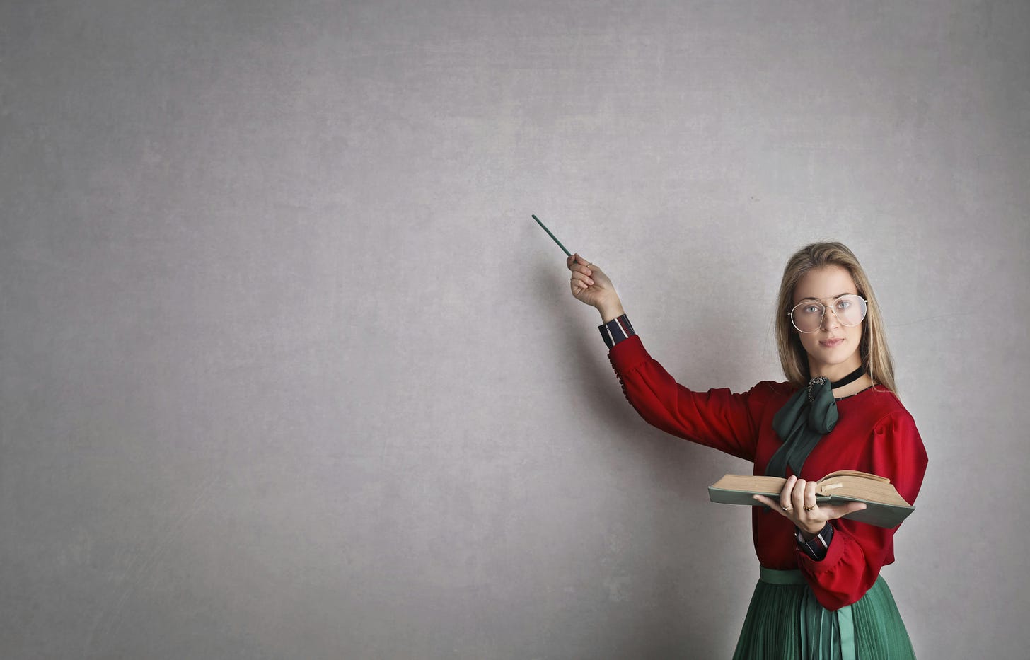 photo entitled "Woman in Discussing A Lesson Plan." she is dressed conservatively, holding a large open book in her left hand. she is holding a pointer in her right hand, which gestures at what appears to be a large blackboard where all the previous writings/markings have been erased.