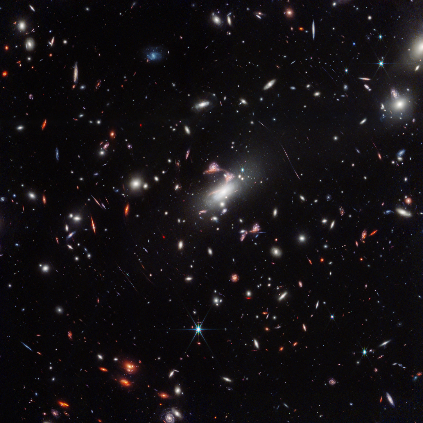 At the center of a field of many colorful galaxies, three elongated red galaxies curve around to approximate the shape of the top of a question mark. Another reddish galaxy appears in about the right position to be the dot of the question mark. A bright white, oval foreground galaxy draws the eye from its position right next to the question mark shape.