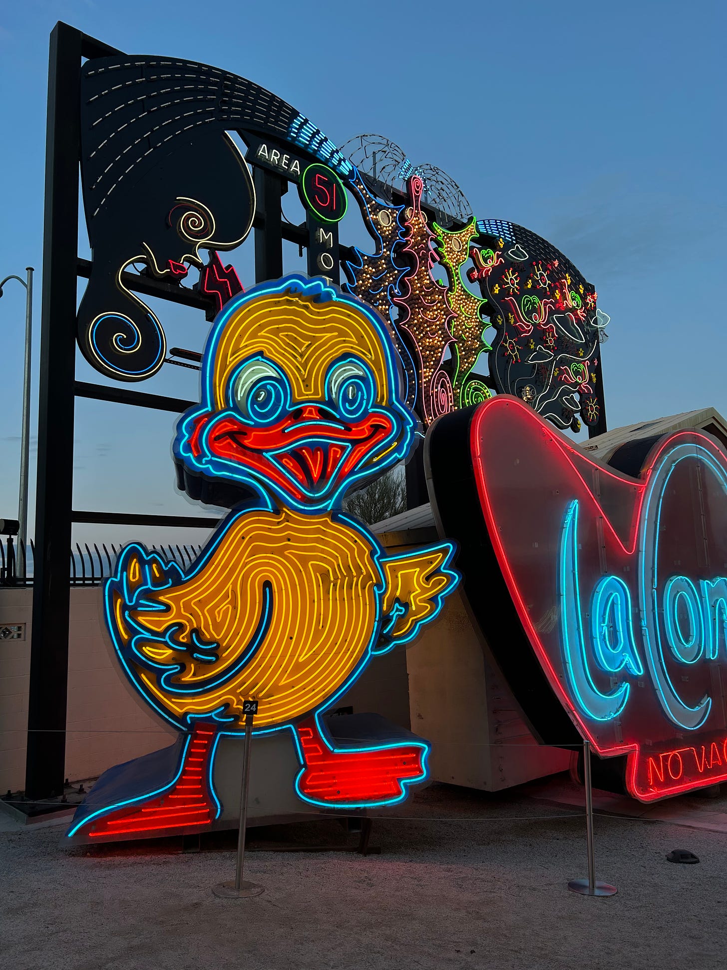 A giant neon sign in the shape of a duck, colored blue, red, and yellow, stands in front of a metal structure with various neon signs tacked onto it; there is also a big red heart-shaped neon sign beside the duck