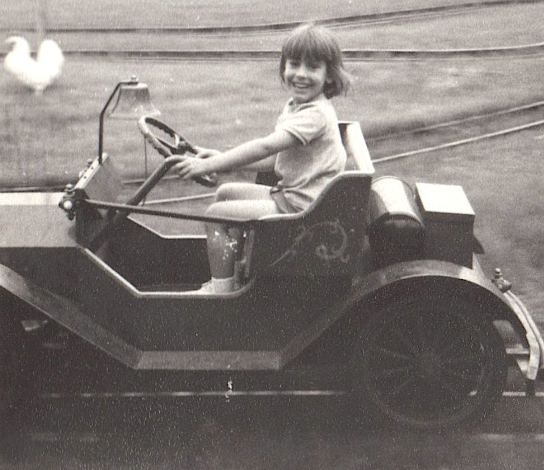 Black-and-white photo of a young girl smiling while looking at the camera and “driving” an open-air car (actually a ride at an amusement park). She has shoulder length light-colored hair with bangs and is wearing shorts and a short-sleeved shirt.