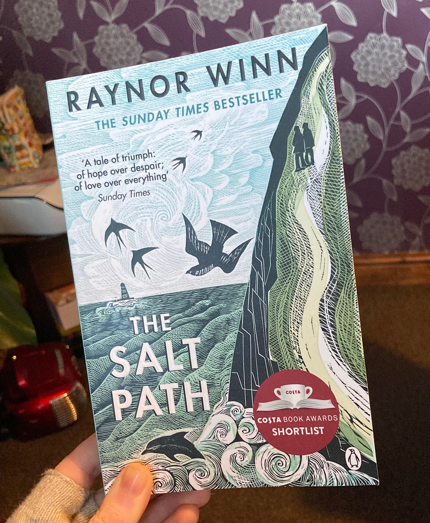 The photo shows a copy of The Salt Path by Raynor Winn, which is being held by Becky.