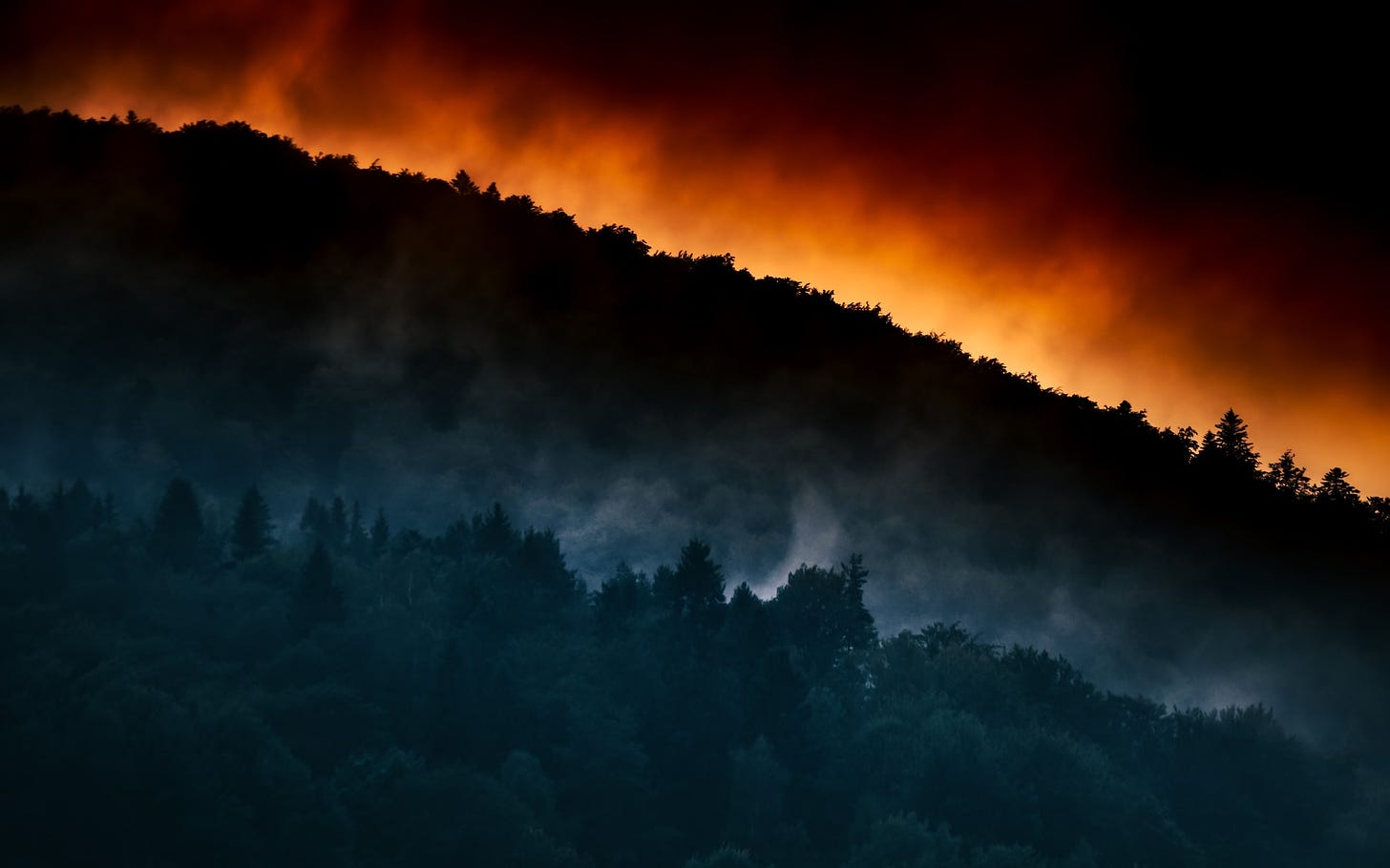 A forested hillside shrouded in smoke with red light peaking over the horizon