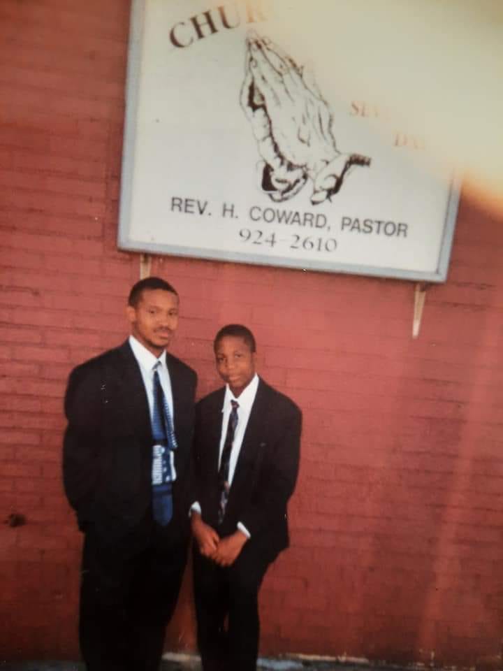 Two young black boys standing in front of a red wall with a sign for the church behind them on the wall wearing dark black suits and white shirts and dark ties.