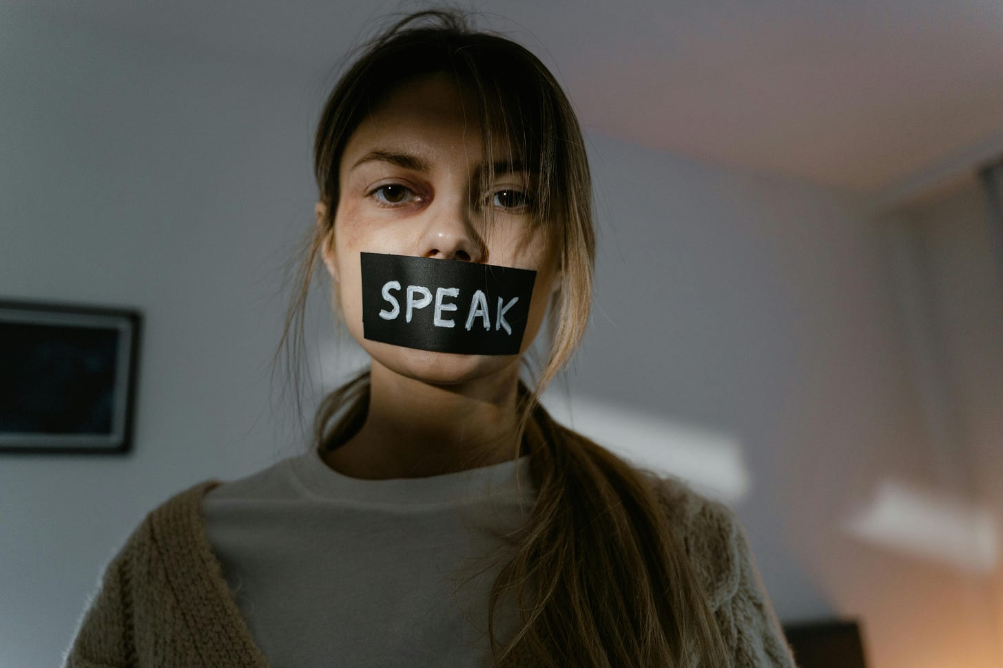 A long haired, slender, figure, appears in a dark room, bruised, with tape over their mouth with the word “speak” written on it. 