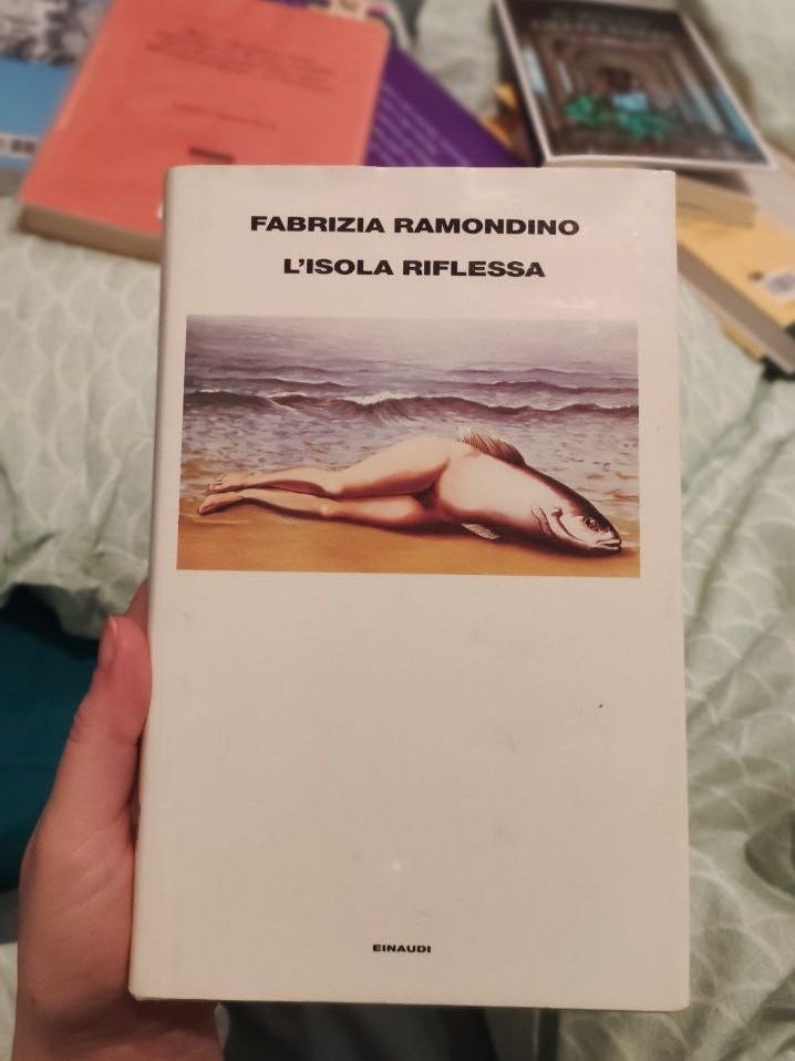Libro bianco con il quadro di Magritte che raffigura un corpo, metà donna e metà pesce, disteso su una spiaggia. Dietro al libro si intravede una coperta e vari libri sparsi