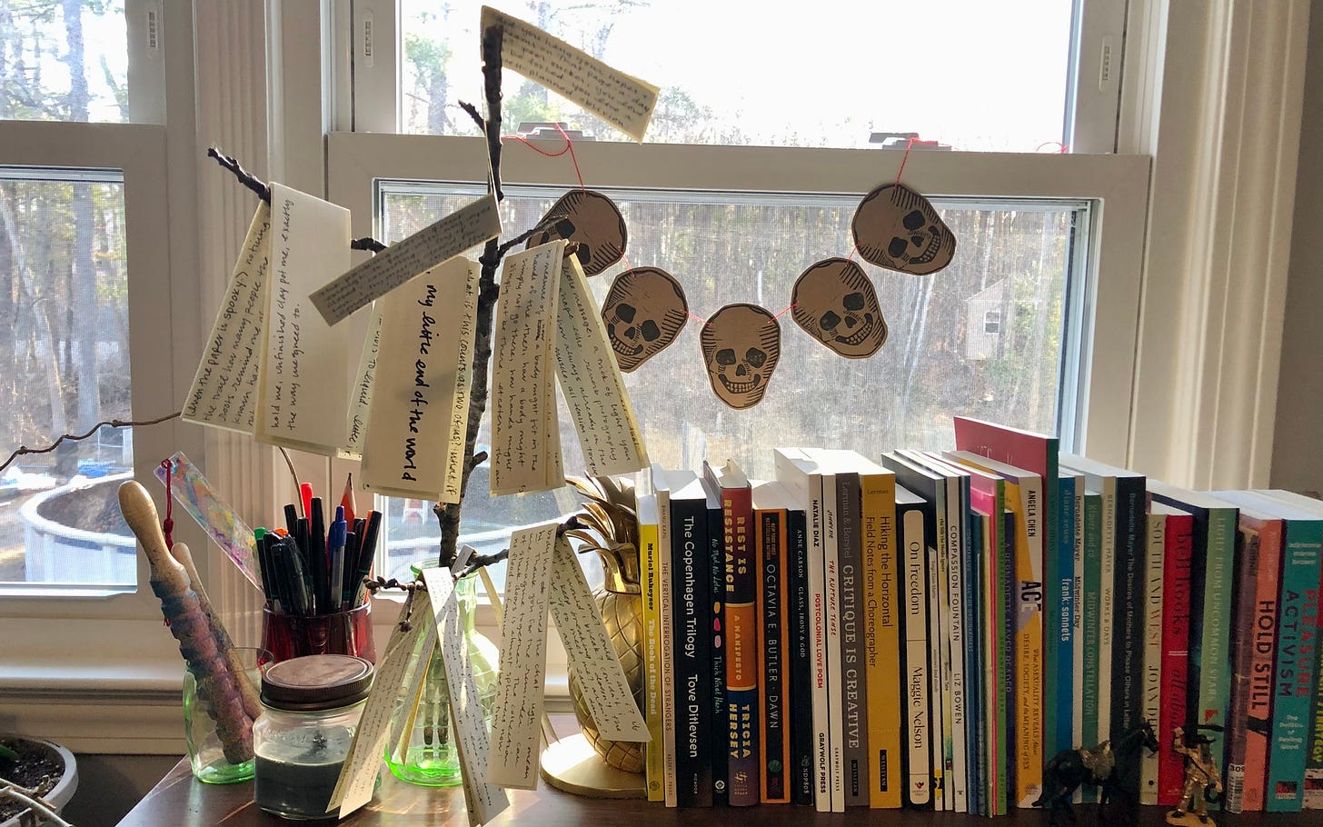 paper scraps, folded and written on in black ink, hung on a small tree branch balanced in a green glass vase. this sits beside a collection of books in front of a sunny window. a garland of skulls hangs in the window. out the windows, trees are visible. in the trees, a treehouse.