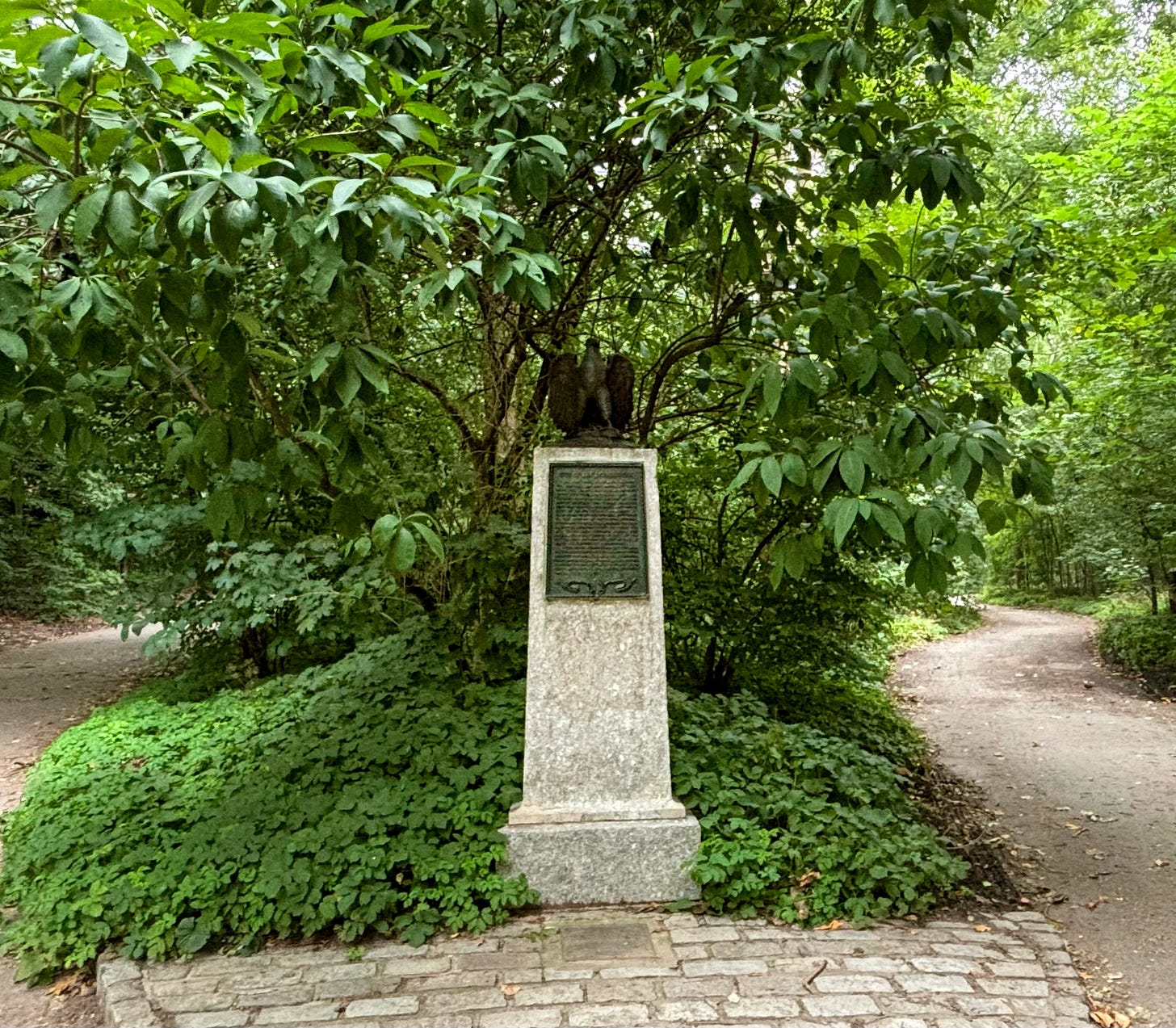 A stone pedestal sits at a fork in the road. A metal eagle perches on top of it and a plaque is on the front.