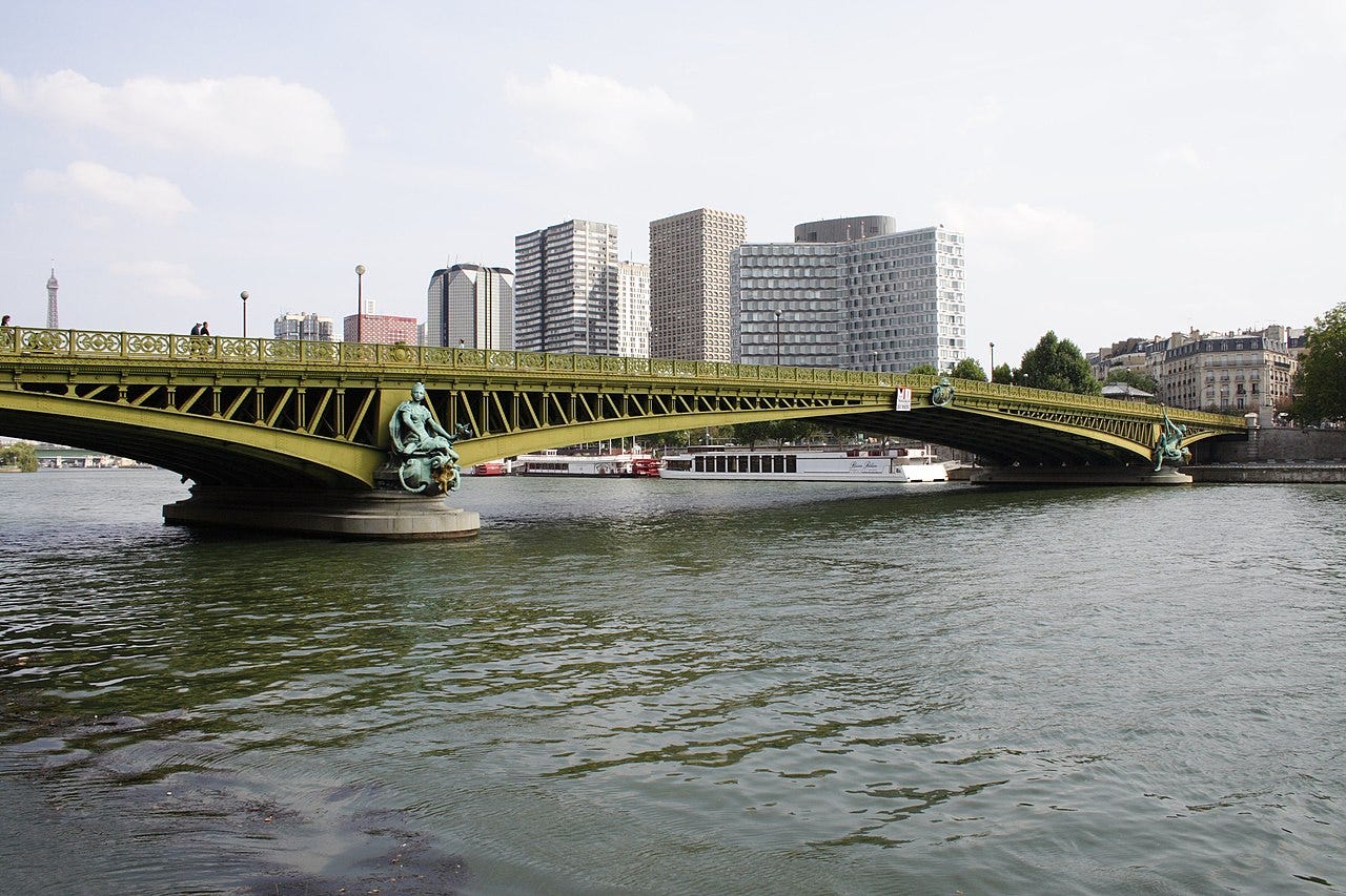 A recent daytime view of the Pont Mirabeau in Paris, a Belle-Epoque bridge with allegorical figures, pictured with modern high-rise buildings visible behind