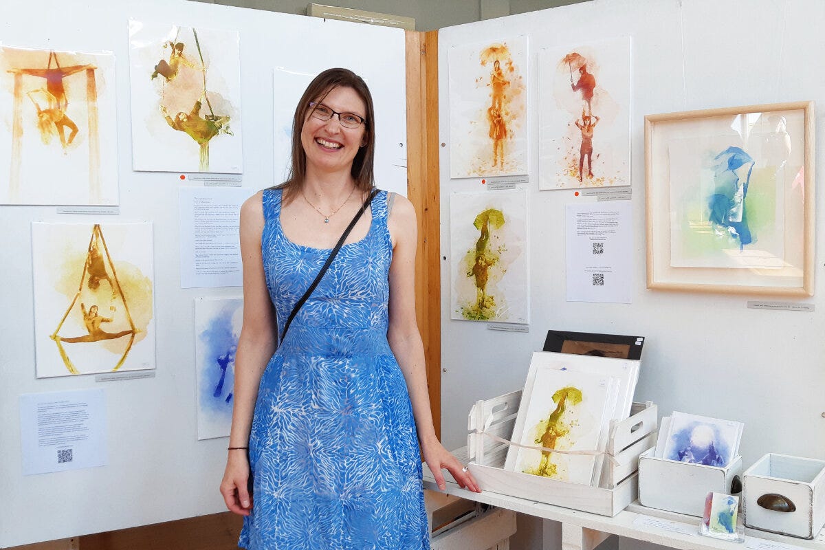 At The Hatch exhibition, Raglan Arts Weekend. Artist, Ailene Cuthbertson is dressed in a blue, summer dress, and stands, smiling, in front of her exhibit of paintings.