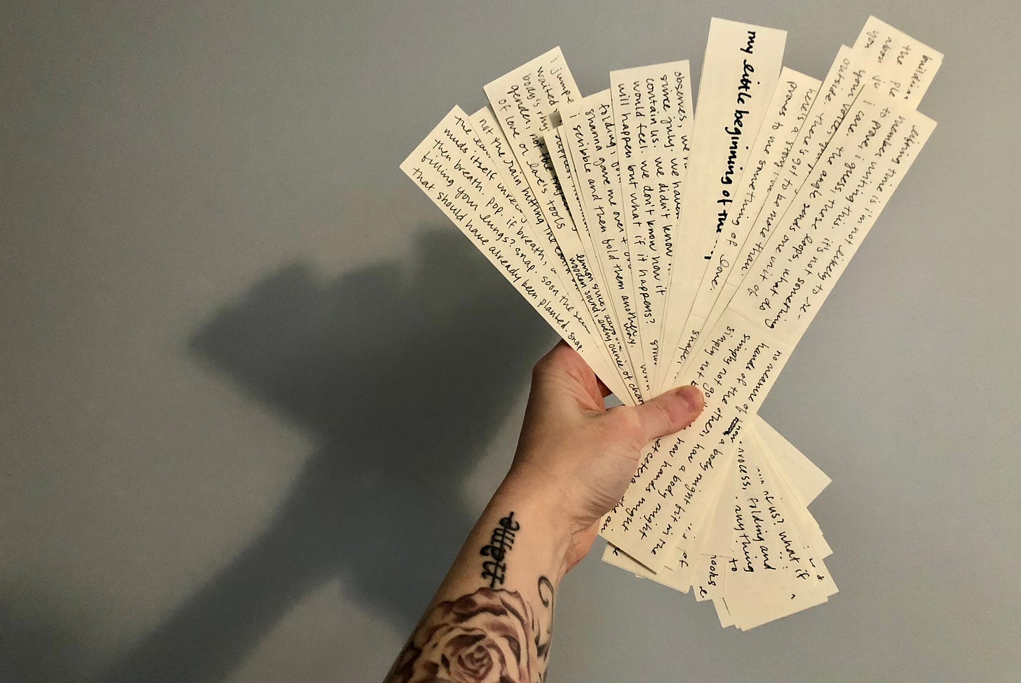a bundle of paper scraps, written on in black ink, fanned out and held up by a tattooed arm. the bundle and the arm cast a shadow on the wall.