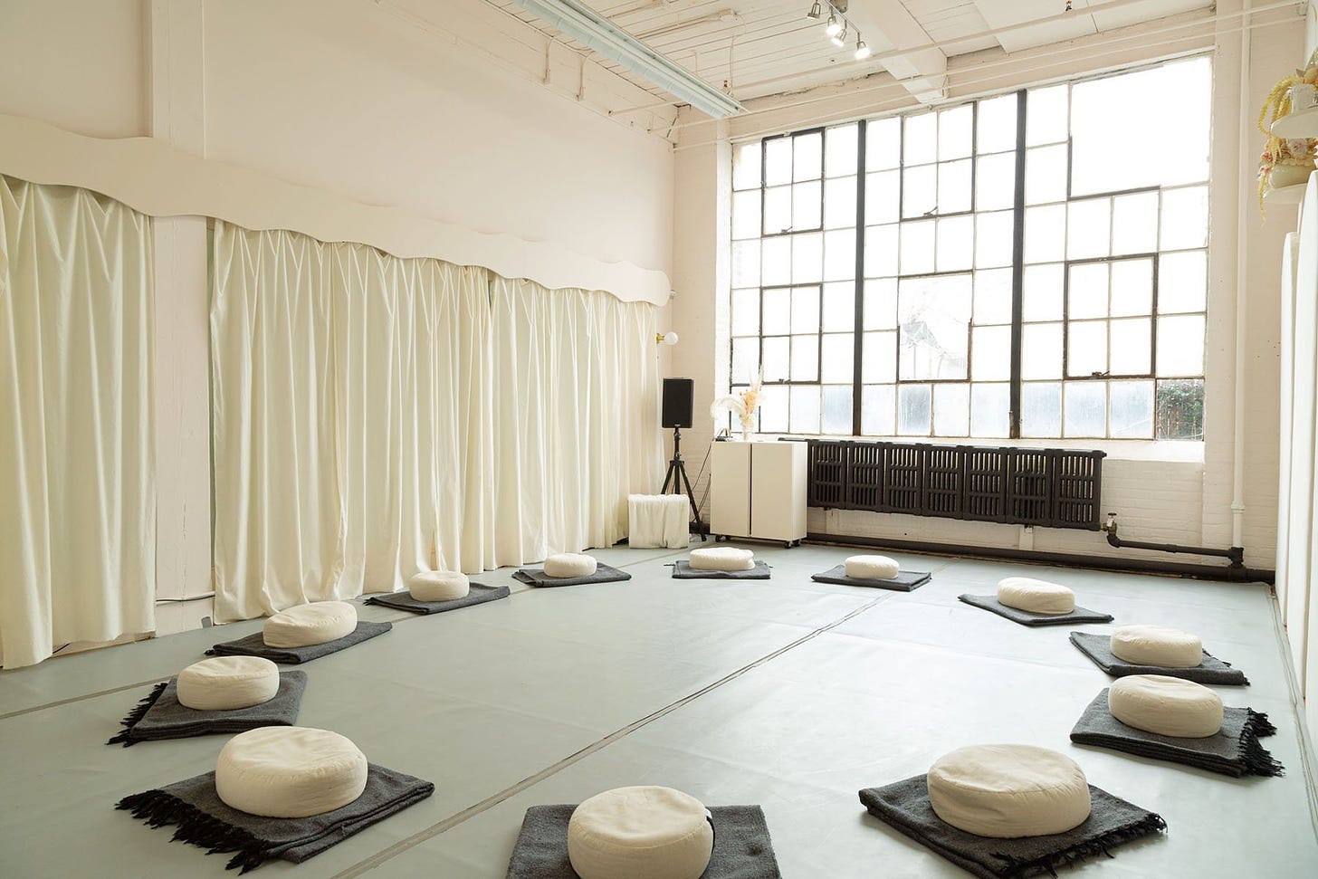 A large movement studio with white walls and gray floors. There's a speaker in the corner and a circle of cushions to sit on. The space has large windows up to the ceiling, letting natural light in.
