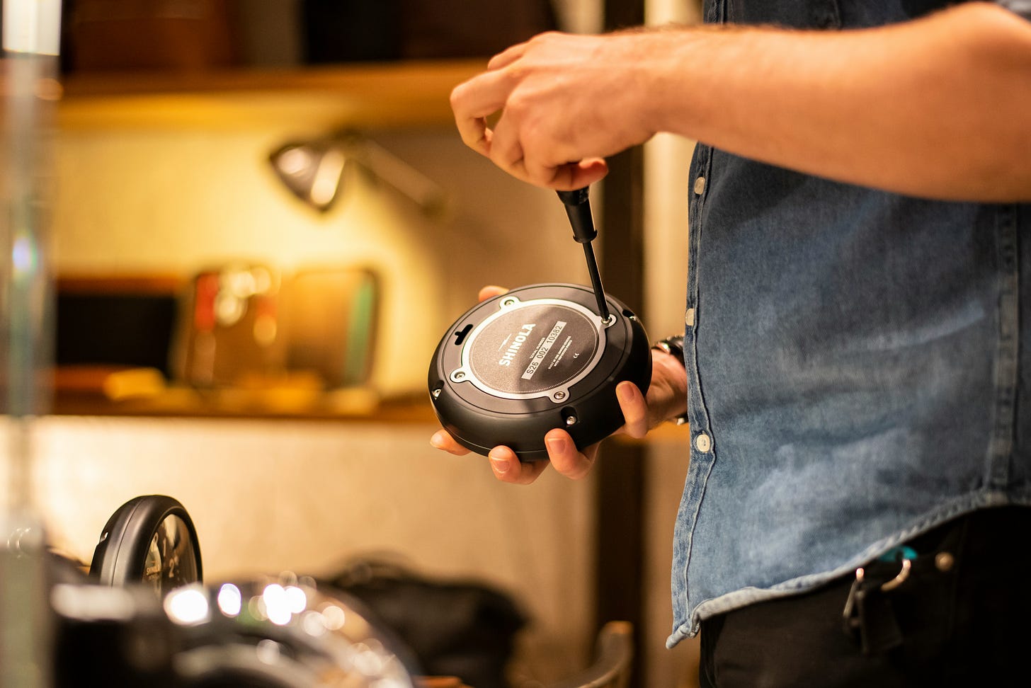 a person holds electronics in one hand while turning a screw with a screwdriver in the other hand. the electronic device is a black disc. only the person's torso and arm are visible. they are wearing a blue demin shirt