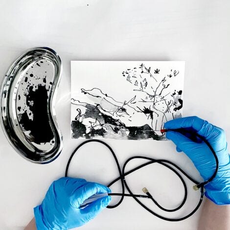 A photo showing a close, bird’s eye view of Sarah’s hands wearing blue medical gloves, resting on a clean white tabletop holding an IV infusion tube needle. The infusion tube is filled with black sumi ink, and Sarah is drawing with the IV needle and the ink on a sheet of paper. Sitting beside the drawing is a stainless-steel kidney shaped surgical dish, splattered with black ink.