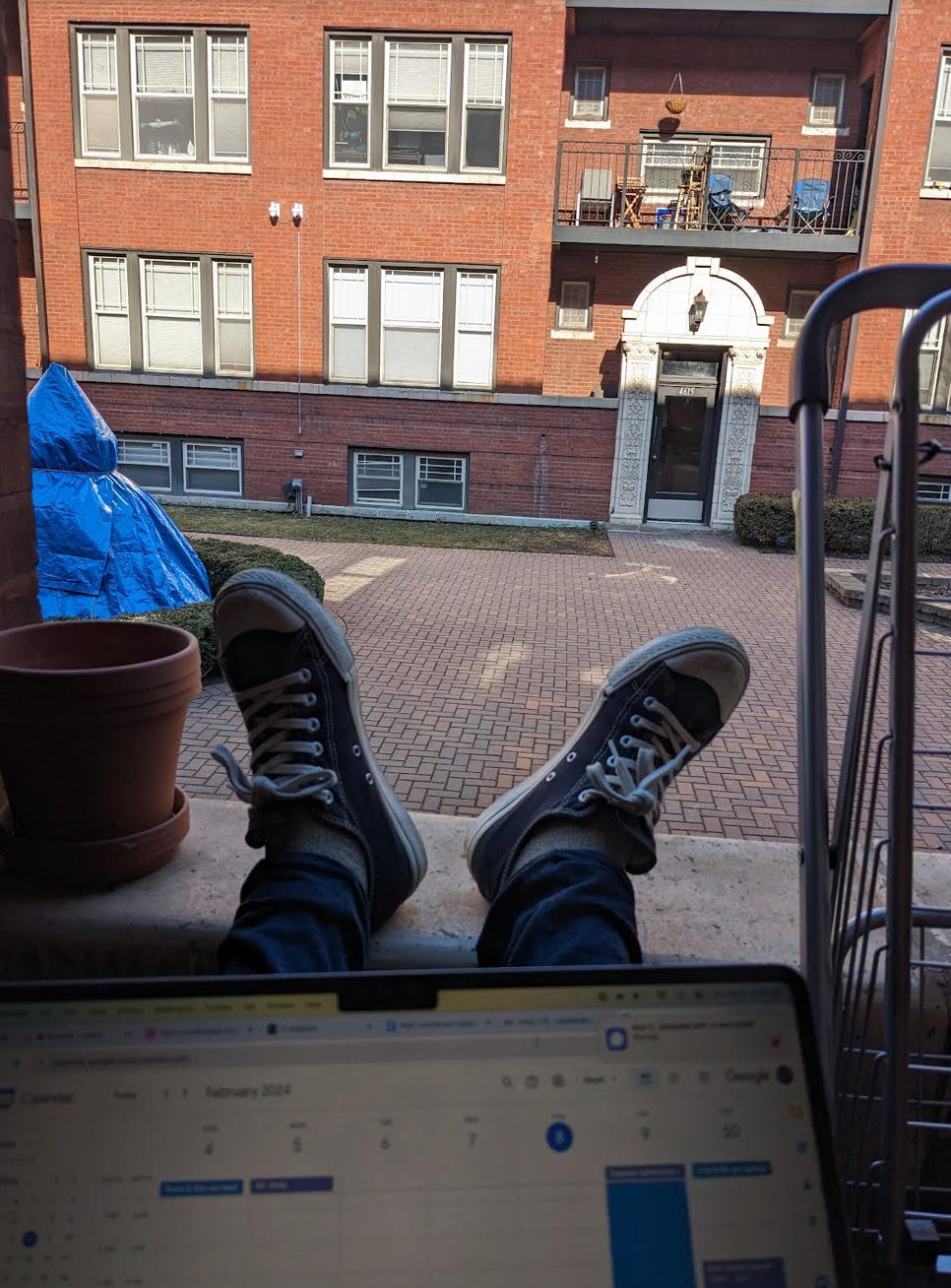 A laptop is on Nathalie's lap as she overlooks the view from her balcony. Her feet are propped up on the balcony sill next to an empty plant pot. Her view is of a red apartment building with an exposed balcony on the top floor 