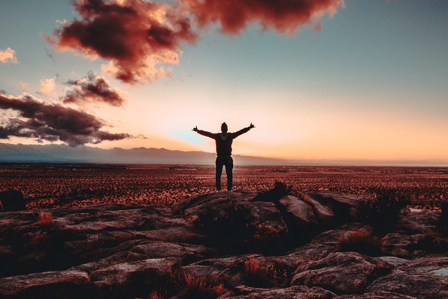 The silhoutette of a man standing with arms outstretched, standing on a rock. In the background is a peach-colored sunrise amidst a dark blue sky. 