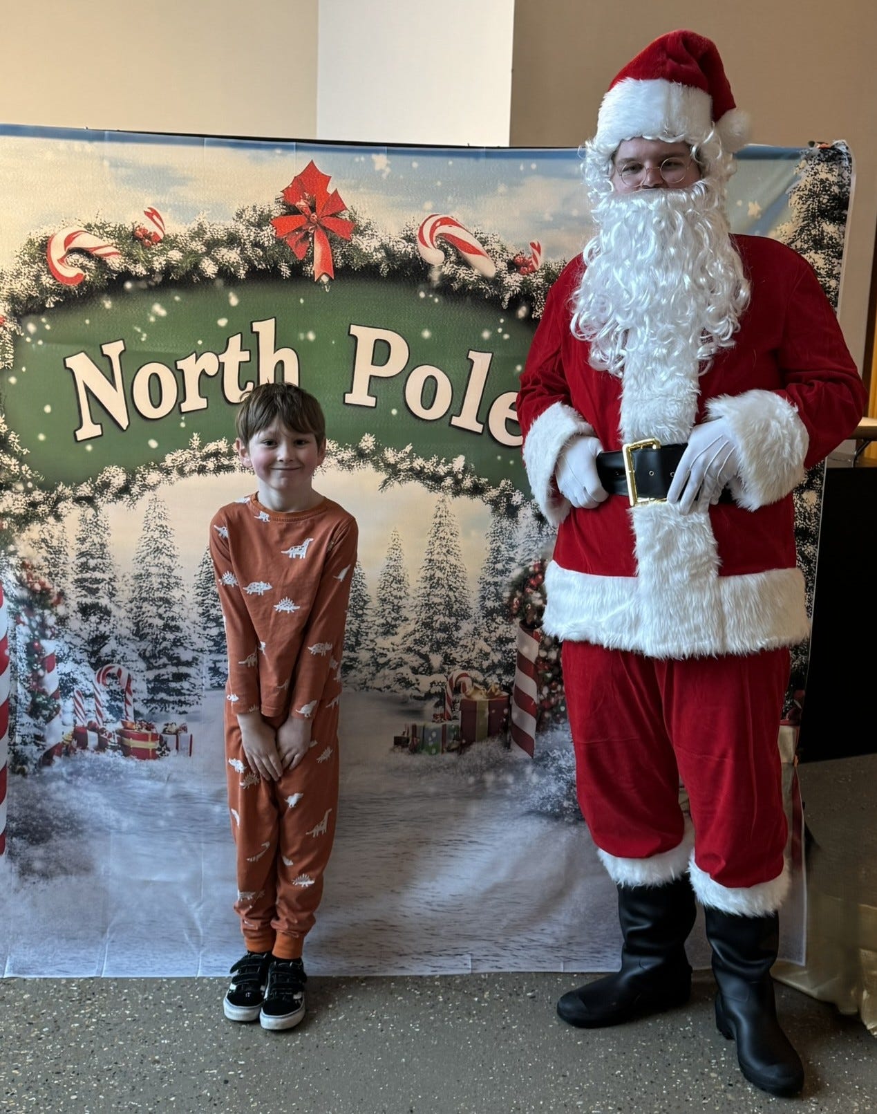 A boy in dinosaur pyjamas is stood in front of a North Pole screen with Father Christmas to his left.  The boys is very excited and shy standing a fair distance away from Santa.