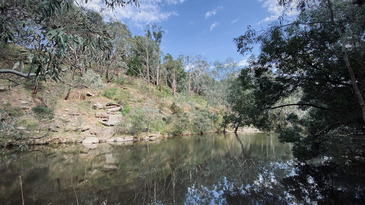 Wide pool in a river in the Australian bush; rocky far bank with eucalypts up the slope; sky and trees reflected in the water.