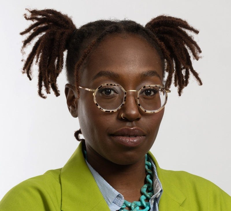 Ariane, young black woman with her hair in ponytails and wearing spotted glasses and a green blazer, looks at the camera
