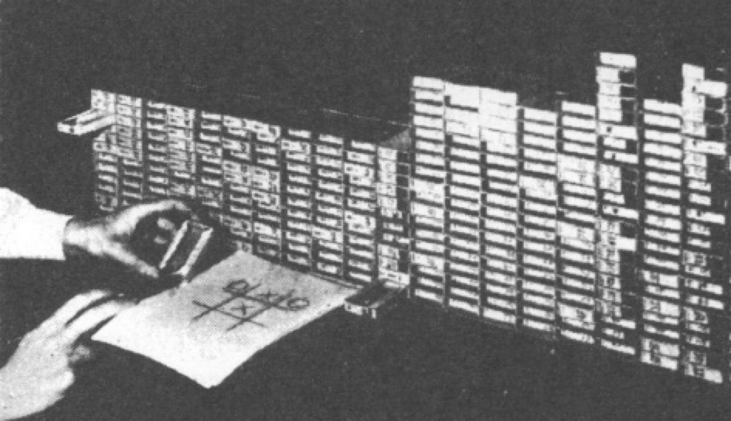 A large bank of matchboxes arranged in columns. In front of the boxes is a partial game of noughts and crosses and a pair of hands.