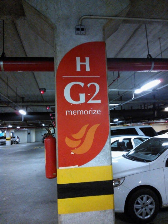 num estacionamento, em destaque uma coluna uma grande plotagem em vermelha com o simbolo do salvador shopping em laranja e escrito com letras brancas e em fonte bem grande: H, G2, memorize