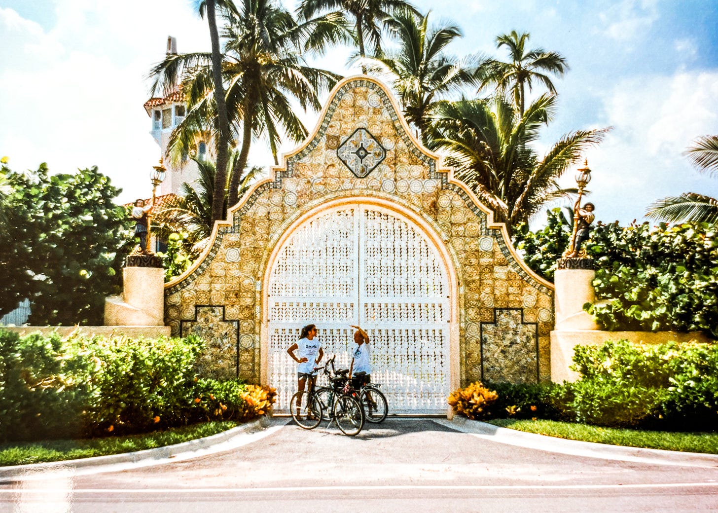 Two people with bicycles standing in front of ornate tile and carved wood gate with tropical foliage surrounding