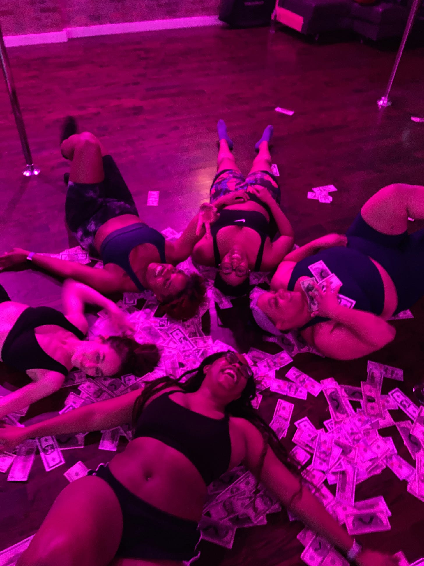 Five women lying on top of fake money on a hardwood floor at a pole studio.