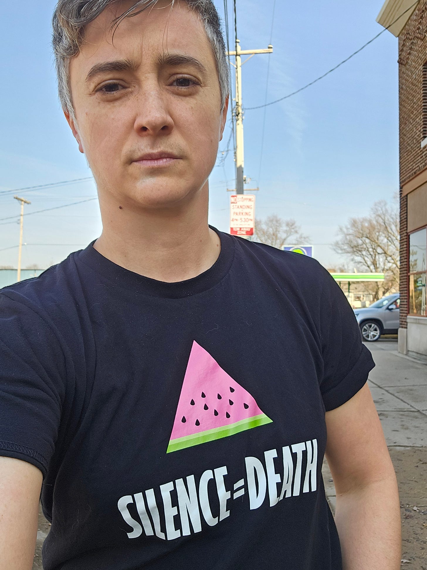 Emily looking into the camera, looking solemn, wears a black t-shirt with the image of a watermelon slice in the shape of the traditional gay pink triangle, and the words "Silence = Death" written below.