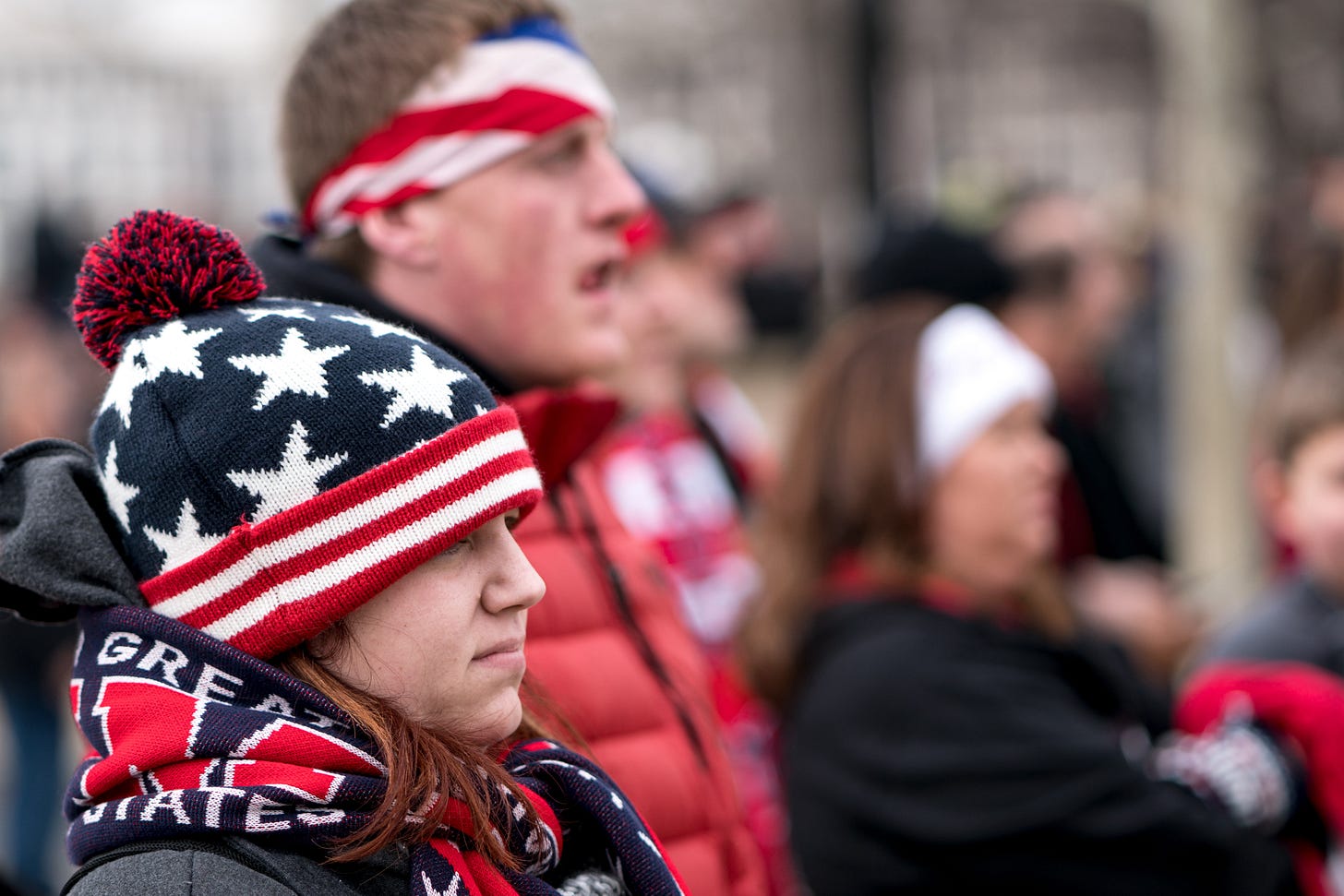 Young woman with woolen stars and stripes hat and scarf that likely reads Make America Great Again (can't read all text on the scarf). Behind her a man with a red, white, blue bandana