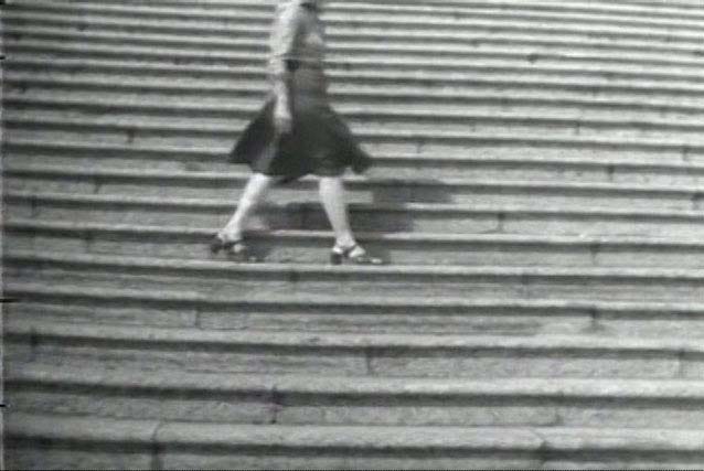 Imagem em preto e branco. Mulher vista do pescoço pra baixo caminhando em uma escadaria.