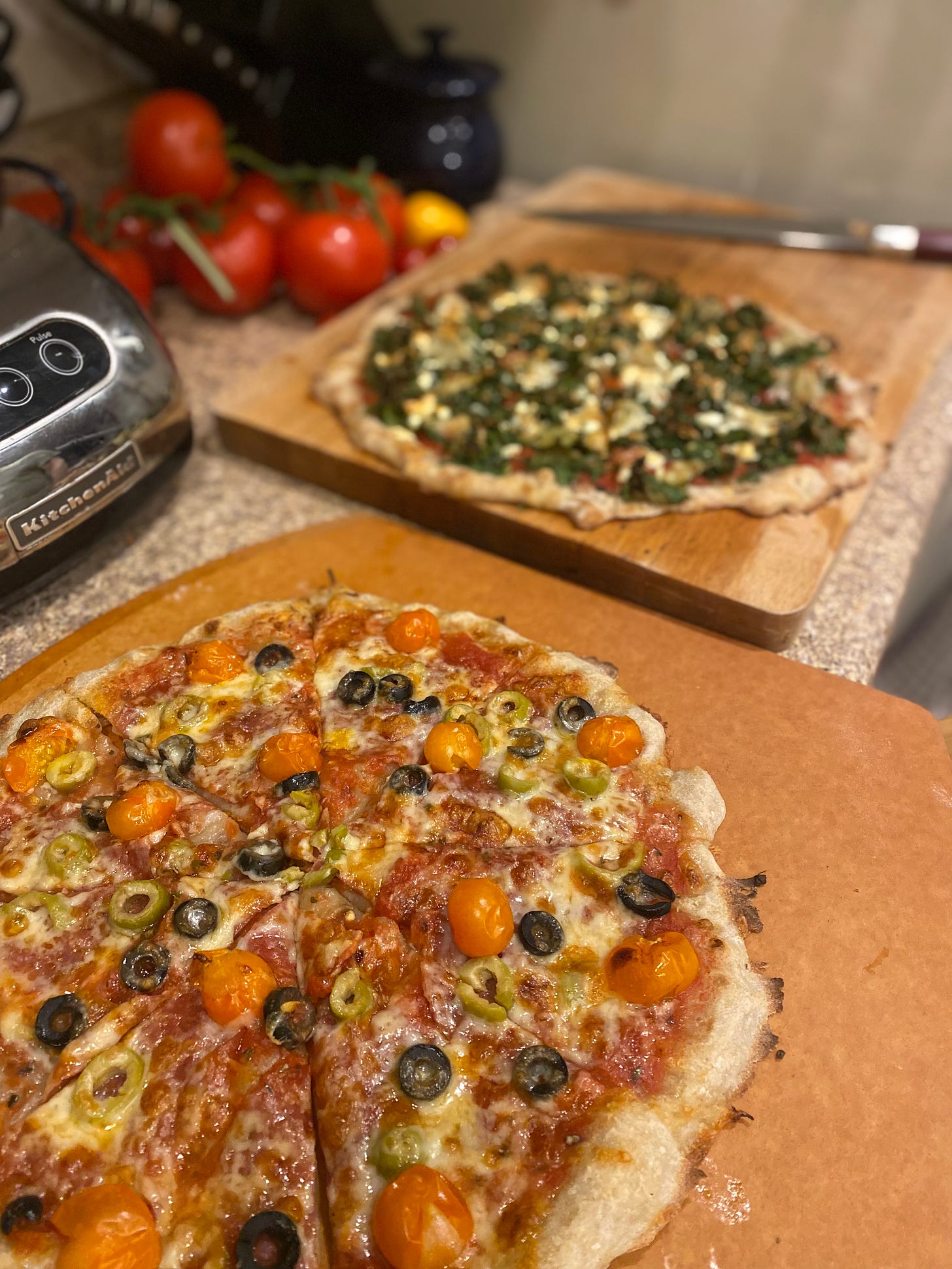 Two pizzas, one on the pizza paddle in the foreground and one on the cutting board in the background. The one in the foreground has salami, sungold tomatoes, and black and green olives. In the background the pizza is blurred, but it is visibly covered in kale and feta.