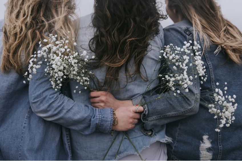 Three girls standing close, holding flowers, seen from the back, like good friends.