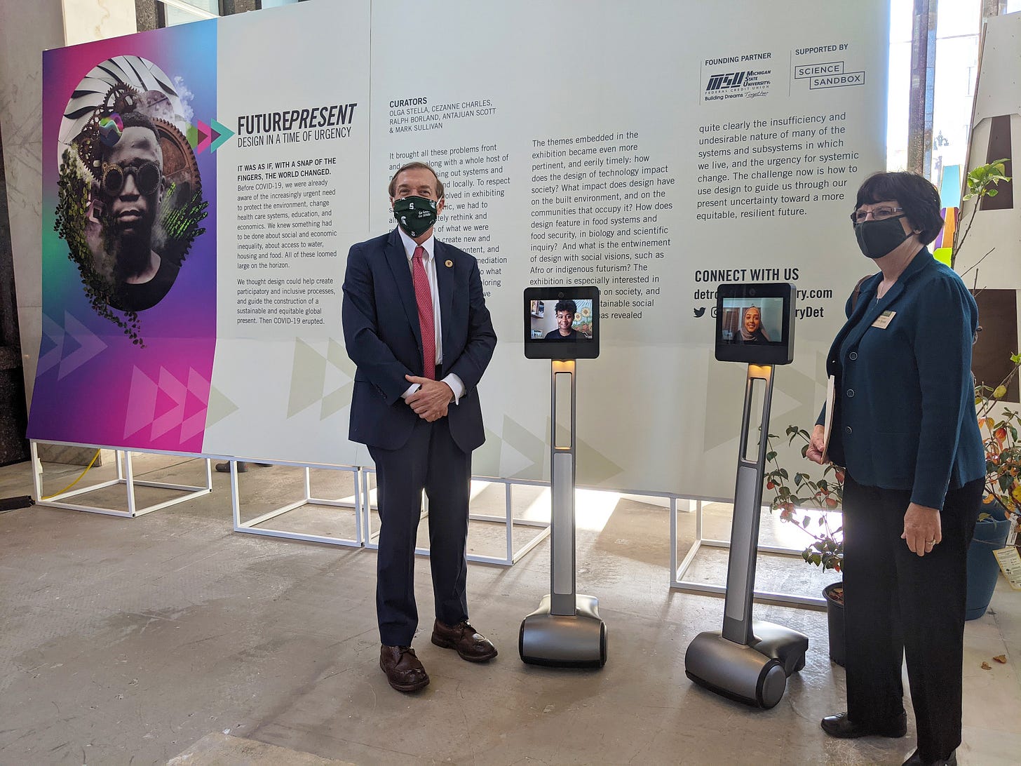 The president and board chair of Michigan State University pose with two telepresence robots in the Future Present exhibition.