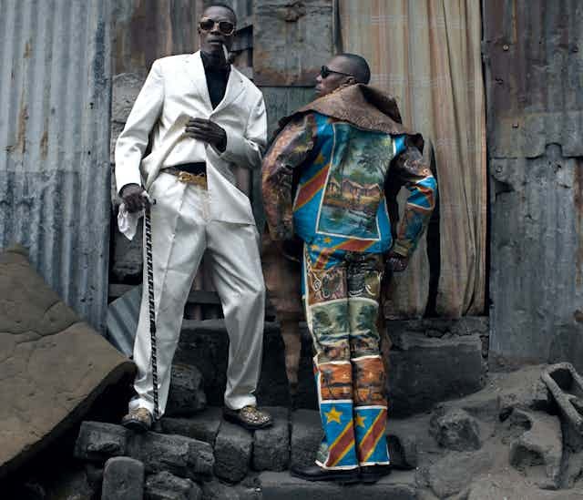 Two men pose in suits against the backdrop of a corrugated iron wall Two men pose in suits against the backdrop of a corrugated iron wall