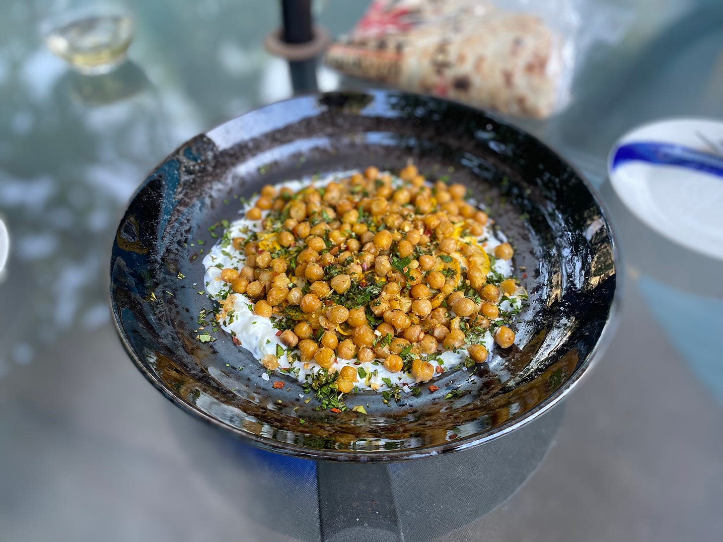 A shallow black bowl of tzatziki and crispy chickpeas sprinkled with herbs and chilies. The yellow zucchini is beneath the chickpeas, but it's almost entirely hidden or blends in with the colour of the chickpeas. In the background is a small white plate with a blue stripe, and a package of taftoon.