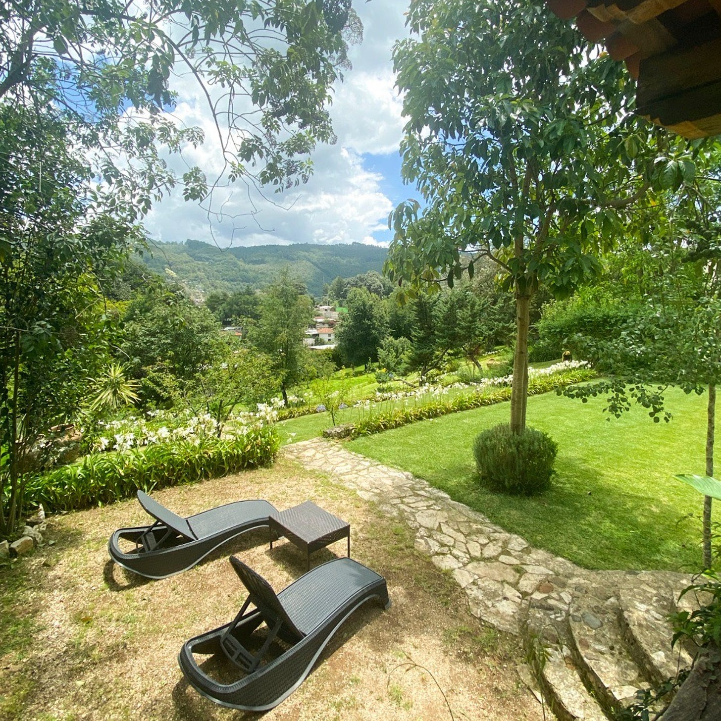 Two outside lounge chairs on grass near stone steps and a walkway outside in the green, wooded hillside of Chiapas in Mexico. The sky is blue with some fluffy white clouds and green mountain in the distance.